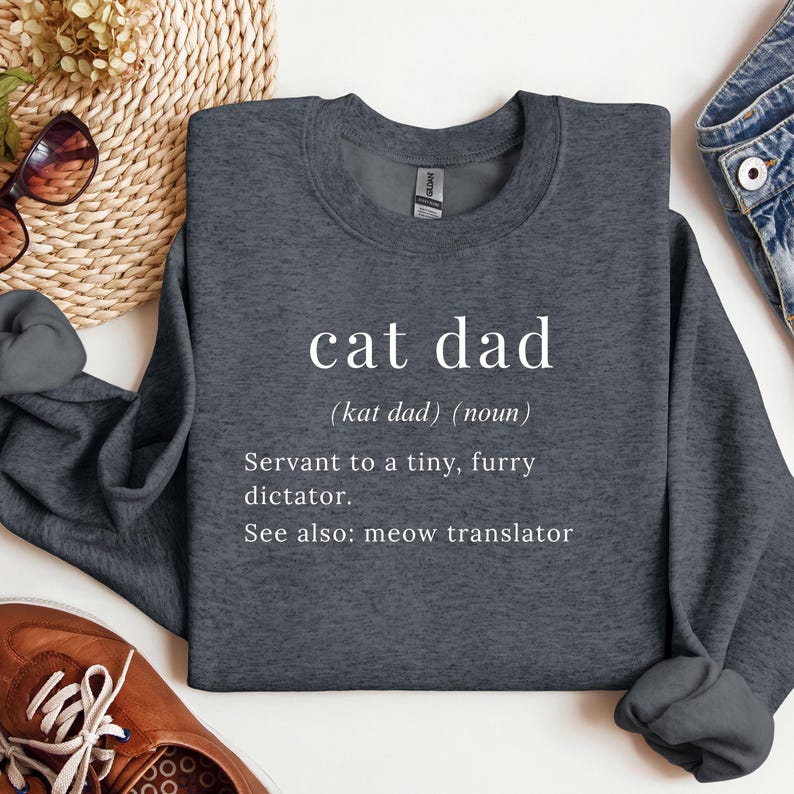 Może przedstawiać: Ciemnoszara bluza z białym napisem "cat dad" i definicją. Definicja brzmi: "Sługa małego, futrzanego dyktatora. Zobacz też: tłumacz miau."
