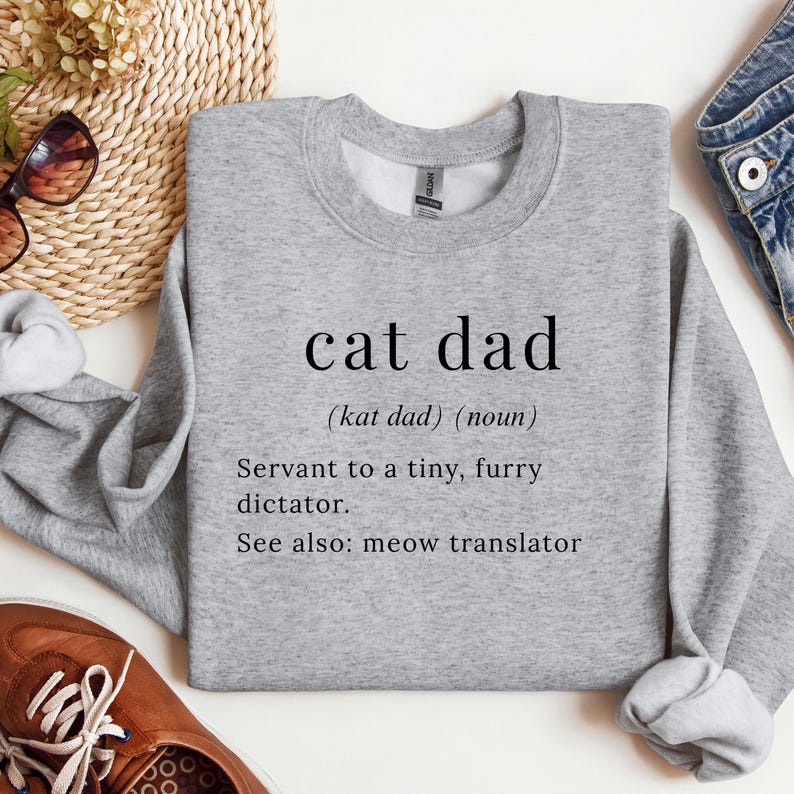 Może przedstawiać: Szara bluza z napisem "cat dad" w czarnym kolorze. Definicja zawiera "Sługa małego, futrzanego dyktatora" oraz "Zobacz też: tłumacz miau".