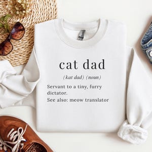 Może przedstawiać: Biała bluza z napisem "cat dad" w czarnym, bezszeryfowym kroju pisma. Poniżej definicja: "(kat dad) (rzeczownik) Sługa małego, włochatego dyktatora. Zobacz też: tłumacz miau."
