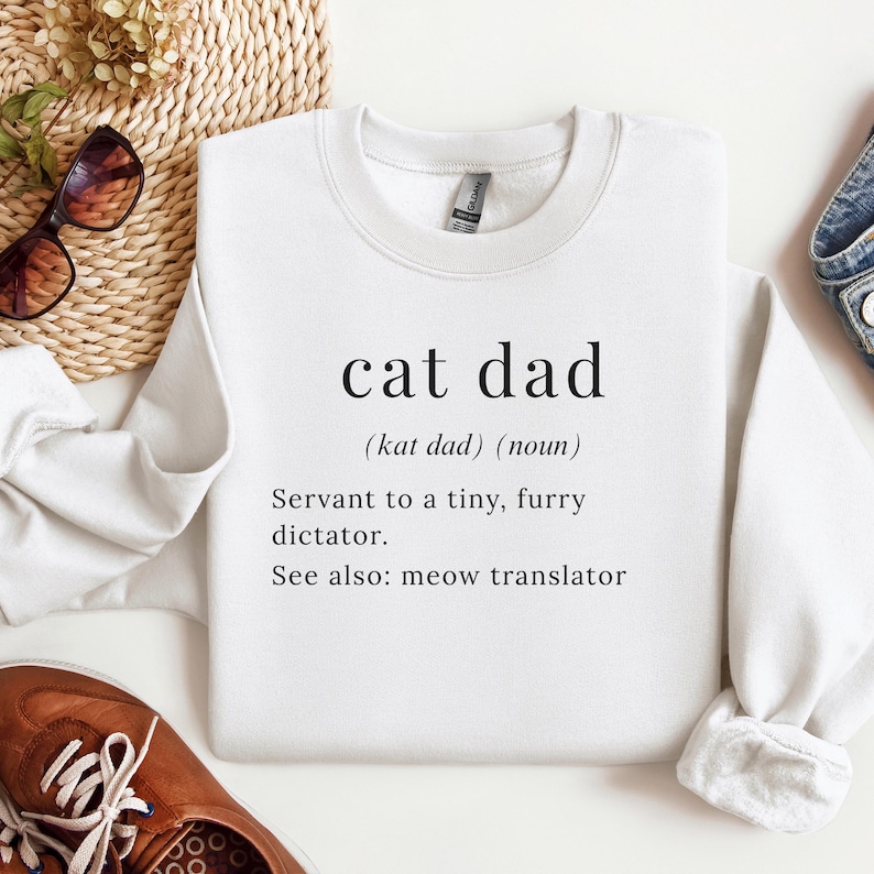 Może przedstawiać: Biała bluza z napisem "cat dad" w czarnym, bezszeryfowym kroju pisma. Poniżej definicja: "(kat dad) (rzeczownik) Sługa małego, włochatego dyktatora. Zobacz też: tłumacz miau."