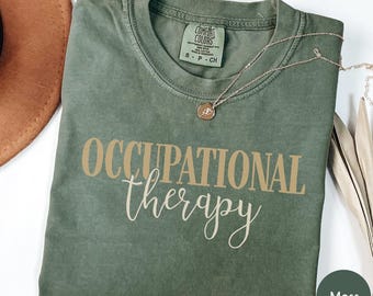 Może przedstawiać: Koszulka z krótkim rękawem w kolorze mchu z napisem OCCUPATIONAL therapy w kolorze beżowym i kremowym. Koszulka posiada metkę marki Comfort Colors przy wewnętrznym dekolcie i jest prezentowana z delikatnym złotym naszyjnikiem.
