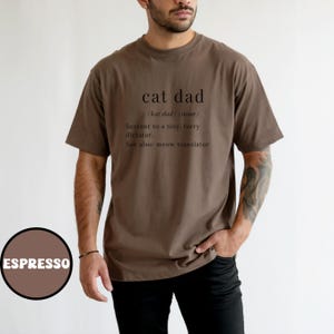 Może przedstawiać: Koszulka w kolorze espresso z napisem "cat dad" i jego definicją: "Sługa małego, futrzanego dyktatora". Koszulka zawiera r&oacute;wnież "Zobacz też: tłumacz miau". Brązowe koło ze słowem "ESPRESSO" znajduje się w lewym dolnym rogu.
