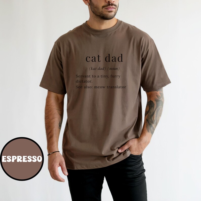 Może przedstawiać: Koszulka w kolorze espresso z napisem "cat dad" i jego definicją: "Sługa małego, futrzanego dyktatora". Koszulka zawiera r&oacute;wnież "Zobacz też: tłumacz miau". Brązowe koło ze słowem "ESPRESSO" znajduje się w lewym dolnym rogu.
