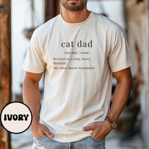 Może przedstawiać: Kremowa koszulka z napisem "cat dad" i jego definicją: "Sługa małego, futrzanego dyktatora. Zobacz też: tłumacz miau." Koszulka to element odzieży codziennej.