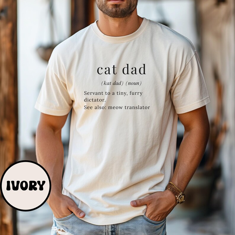 Może przedstawiać: Kremowa koszulka z napisem "cat dad" i jego definicją: "Sługa małego, futrzanego dyktatora. Zobacz też: tłumacz miau." Koszulka to element odzieży codziennej.