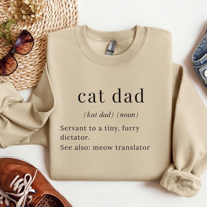 Może przedstawiać: Beżowa bluza z napisem "cat dad" pogrubioną czcionką. Poniżej definicja: "(kat dad) (rzeczownik) Sługa małego, futrzanego dyktatora. Zobacz też: tłumacz miau."