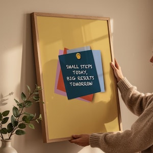 May include: A framed poster with a yellow background and a wooden frame. The poster has a blue sticky note with the text "SMALL STEPS TODAY, BIG RESULTS TOMORROW". A woman is holding the poster.