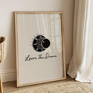 May include: A framed print with a beige background features a black and white graphic of a dial with the text "Lower The Drama" in a script font. The print is in a light wood frame and is leaning against a wall.