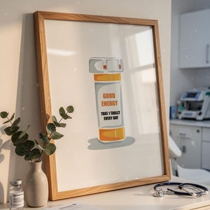 Op de afbeelding: Een ingelijste print met een cartoon pillenfles met de tekst "GOOD ENERGY" en "TAKE 1 TABLET EVERY DAY". De fles is oranje, wit en grijs. Een stethoscoop en een kleine vaas met groen staan op de voorgrond.