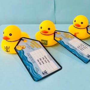 May include: Three yellow rubber duck toys with orange beaks and black eyes are displayed with luggage tags. The tags have a blue wave design and the text "Commemorative Duck". The background is a light blue surface.