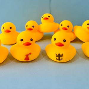 May include: A collection of yellow rubber duck toys with black eyes and orange beaks. Some ducks have logos, including a cruise line logo, a windmill, and a Mickey Mouse silhouette. The ducks are arranged on a light blue surface.