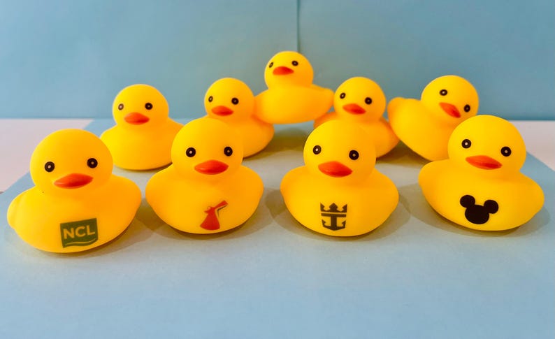 May include: A collection of yellow rubber duck toys with black eyes and orange beaks. Some ducks have logos, including a cruise line logo, a windmill, and a Mickey Mouse silhouette. The ducks are arranged on a light blue surface.