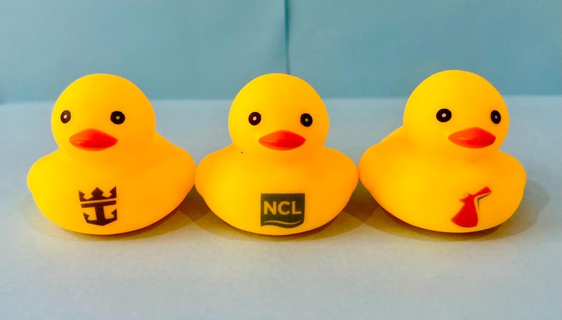 May include: Three yellow rubber duck toys with black eyes and orange beaks. Each duck has a different logo on its chest: a crown, the letters "NCL" in a rectangle, and a red and white logo.