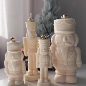 Könnte beinhalten: Vier weiße Nussknacker-Kerzen mit goldenen Akzenten. Die Kerzen sind in verschiedenen Größen und in einer Reihe auf einer weißen Oberfläche angeordnet.