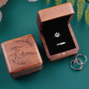 May include: Two wooden ring boxes, one open with two rings and one closed with text. The open box has a black interior. The closed box has the text "STEVE & WANDA The Williams May 15, 2023". Two additional rings are visible.