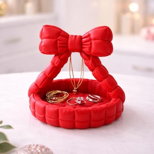 May include: A red jewelry tray shaped like a bow, holding gold bracelets, rings, and a necklace. The tray has a quilted texture and is designed to hold jewelry. The background is blurred, with soft lighting.
