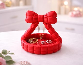 Può includere: Un vassoio portagioie rosso a forma di fiocco, con bracciali, anelli e una collana d'oro. Il vassoio ha una texture trapuntata ed è progettato per contenere gioielli. Lo sfondo è sfocato, con un'illuminazione soffusa.