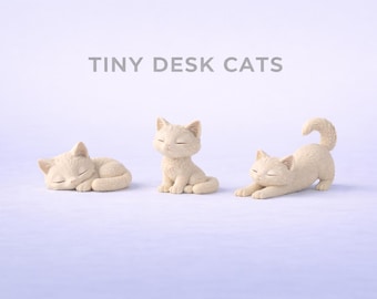 May include: Three small, light beige cat figurines arranged in a row against a light background. The text TINY DESK CATS is printed above them. One cat is curled up sleeping, one is sitting upright, and one is stretching with its tail raised.