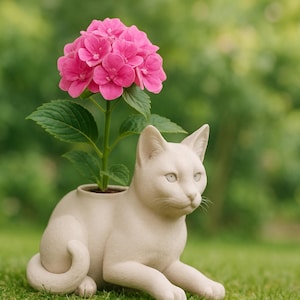 May include: A white ceramic cat planter with a pink hydrangea plant growing out of the top. The cat planter is sitting on green grass.