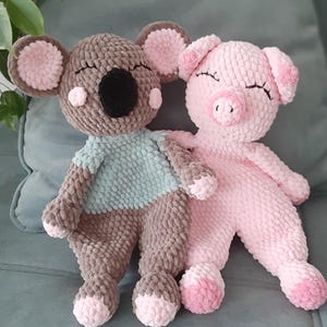 May include: Two handmade plush toys: a koala and a pig. The koala is gray with pink accents and a blue shirt. The pig is pink with closed eyes. Both toys are made of soft, textured material and appear cuddly.