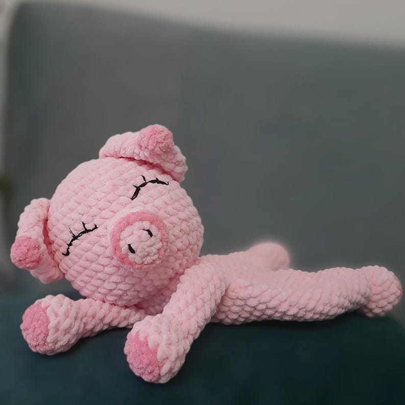May include: A handmade pink crochet pig plush toy. The pig is lying down with its eyes closed, and has a small, round snout, ears, and feet. The plush toy is made of soft, textured yarn.