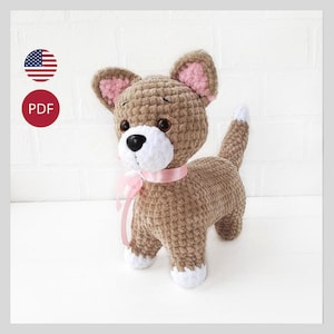 Puede incluir: Patrón de crochet para un juguete de peluche de perro marrón y blanco con orejas rosas y una cinta rosa alrededor del cuello. El patrón está disponible para descargar en PDF.