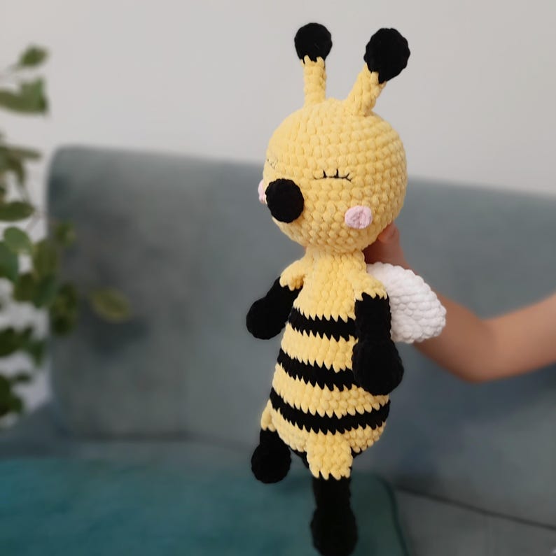 May include: A handmade crochet bee plush toy. The toy is primarily yellow with black stripes, black limbs, and black antennae. It has a sweet face with closed eyes, pink cheeks, and a white wing.