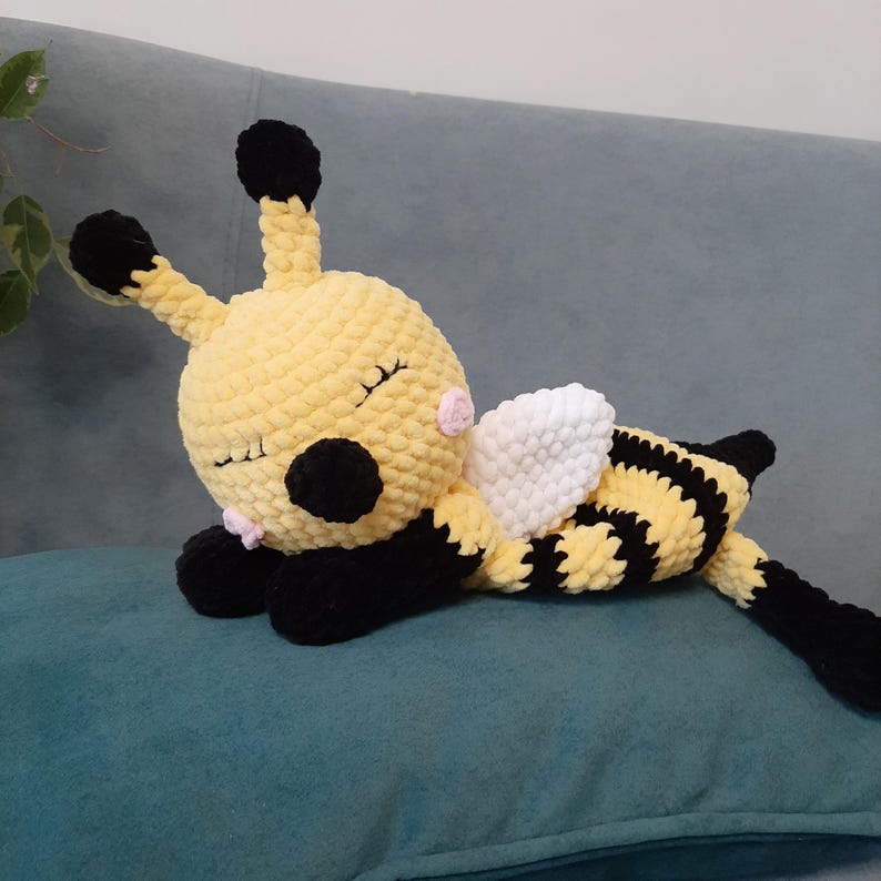 May include: A handmade yellow and black plush bee toy with white wings, black antennae, and closed eyes. The bee is lying on a teal pillow, showcasing a soft, textured design. The toy is a cute, cuddly item.