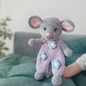 May include: A gray crocheted mouse plush toy with pink ears and blue flower accents. The toy is wearing a purple crocheted outfit.