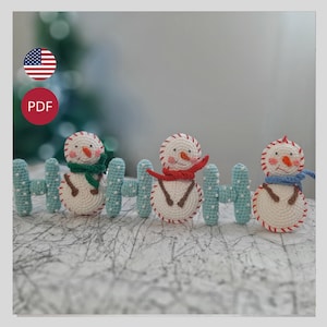 Puede incluir: Tres muñecos de nieve de ganchillo con narices de zanahoria, brazos de ramita y sonrisas cosidas en rojo están detrás de una valla azul claro. Cada muñeco de nieve lleva una bufanda de diferente color: verde, roja y azul.