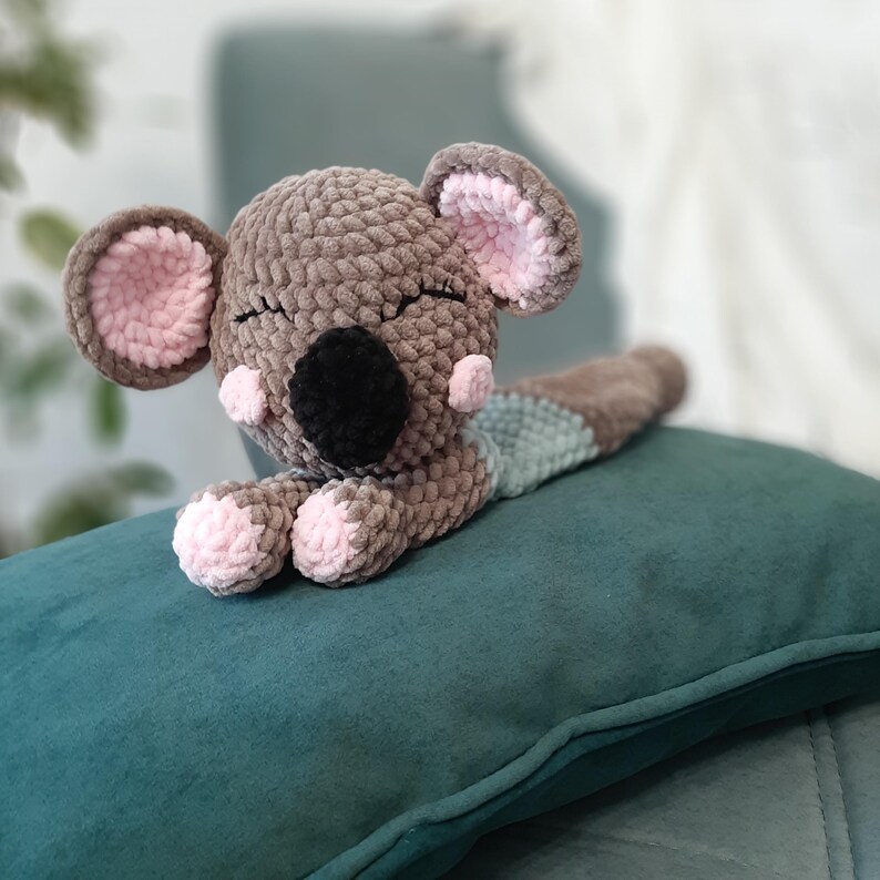 May include: A handmade crochet koala bear in shades of gray, brown, black, and pink, lying on a teal pillow. The koala has closed eyes, a black nose, and pink accents on its ears and paws. The plush toy is a soft, cuddly item.