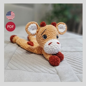 Könnte beinhalten: Ein gehäkeltes Giraffen-Plüschtier mit braunem und weißem Fell, roten Hufen und schwarzen Augen. Die Giraffe liegt auf einer grauen, strukturierten Oberfläche.