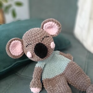 May include: A handmade, plush koala bear in shades of brown, gray, and pink. The koala has a black nose, closed eyes, and a light blue shirt. The ears and paws are accented with pink. The toy is lying on a teal surface.