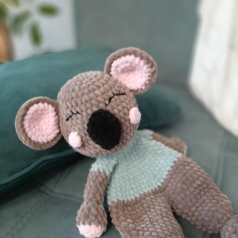 May include: A handmade, plush koala bear in shades of brown, gray, and pink. The koala has a black nose, closed eyes, and a light blue shirt. The ears and paws are accented with pink. The toy is lying on a teal surface.