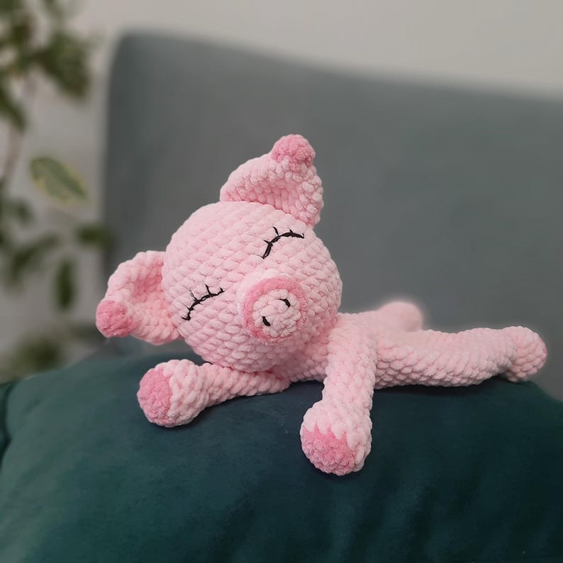 May include: A handmade pink crochet pig plush toy. The pig has black stitched eyes and a curled tail. The pig is lying down on a dark green surface. The pig's ears, snout, and feet are a darker shade of pink.