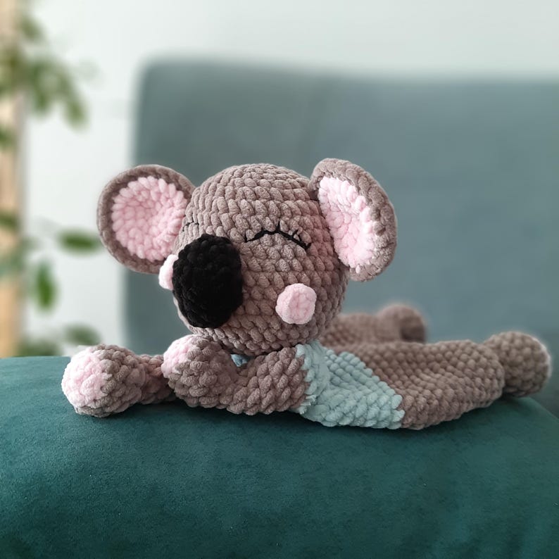 May include: A handmade, plush koala bear in shades of gray, brown, and pink. The koala has a black nose, closed eyes, and pink accents on its ears and cheeks. It is lying on its stomach with a light blue patch on its belly.