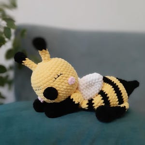 May include: A handmade, plush bee toy in yellow, black, and white. The bee has black and yellow striped body, black antennae, and white wings. The bee is lying down with its eyes closed, suggesting it is sleeping.