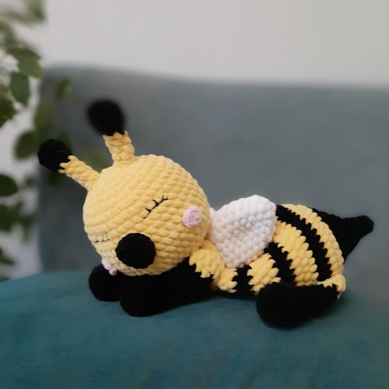 May include: A handmade, plush bee toy in yellow, black, and white. The bee has black and yellow striped body, black antennae, and white wings. The bee is lying down with its eyes closed, suggesting it is sleeping.