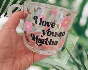 Puede incluir: Taza de vidrio transparente con asa, decorada con flores rosas y el texto negro "I love You so Matcha". La taza tiene una forma curva y se muestra sobre un fondo de follaje verde.
