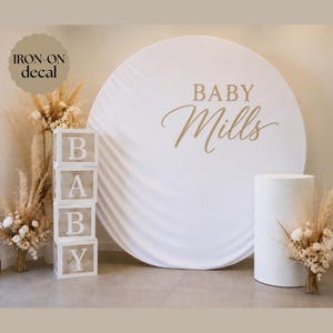 May include: A large white circular backdrop featuring the gold text BABY Mills, accompanied by a vertical stack of four transparent square boxes with white letters spelling BABY and a white cylindrical pedestal, all decorated with dried floral arrangements.
