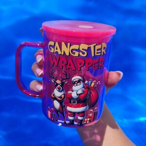 Puede incluir: Un vaso de plástico rosa con tapa transparente y asa. El vaso tiene un gráfico de Santa Claus y un reno con gafas de sol y el texto "Gangster Wrapper".