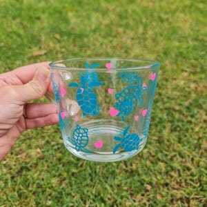 Könnte beinhalten: Transparente Glas-Tasse mit türkisfarbenen Meeresschildkröten und rosa Herzen. Die Tasse hat einen Henkel und steht vor einem Hintergrund aus grünem Gras. Das Design wiederholt sich um die Außenseite der Tasse.