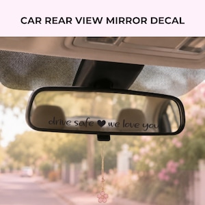 May include: A black vinyl decal applied to a car rear view mirror featuring the text drive safe followed by a heart symbol and the text we love you in a cursive script font. The decal is positioned horizontally across the centre of the mirror.