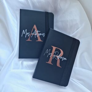 May include: Two black rectangular notebooks with elastic closure bands. Each notebook features a large metallic rose gold initial letter and a white cursive script name printed on the front cover. One reads Mrs Artemis and the other reads Mrs Robinson.