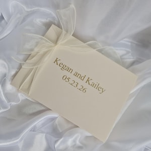 May include: A rectangular cream-coloured guest book featuring the gold-printed text Kegan and Kailey 05.23.26 on the front cover. The book is adorned with a delicate, sheer white organza ribbon tied in a bow at the top left corner of the cover.