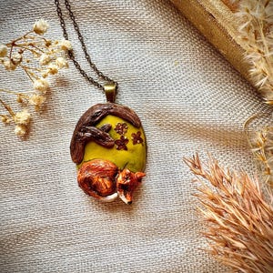 Puede incluir: Un collar con un colgante que representa un zorro durmiendo acurrucado sobre un fondo verde y amarillo con ramas marrones y flores blancas.