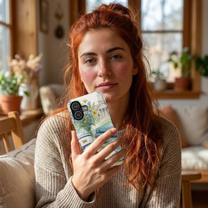 May include: A white phone case featuring a watercolor illustration of a building, flowers, and books. The case has a black camera lens cutout and a blue and yellow color scheme. The word "NURSING" is visible on the case.
