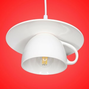 Könnte beinhalten: Eine weiße Hängeleuchte aus Keramik in Form einer Tasse und Untertasse, die an einem weißen Kabel hängt. Die Tasse ist umgedreht und dient als Lampenschirm, wobei eine Glühbirne mit sichtbarem Glühfaden im Inneren der Tasse zu sehen ist.