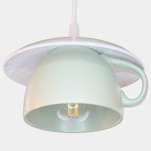 Könnte beinhalten: Eine Pendelleuchte mit einer hellgrünen Keramiktasse als Lampenschirm, die von einer weißen Untertasse gekrönt wird. Die Tasse hat einen seitlichen Henkel und eine einzelne Glühbirne mit sichtbarem Faden an einem weißen Kabel.