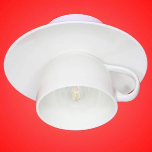 Könnte beinhalten: Eine weiße Teetasse mit Untertasse aus Keramik, die als Deckenleuchte umfunktioniert wurde. Die Tasse ist umgedreht und im Inneren befindet sich eine sichtbare Glühbirne mit Filament. Die Untertasse dient als Deckenplatte mit glatter Oberfläche.
